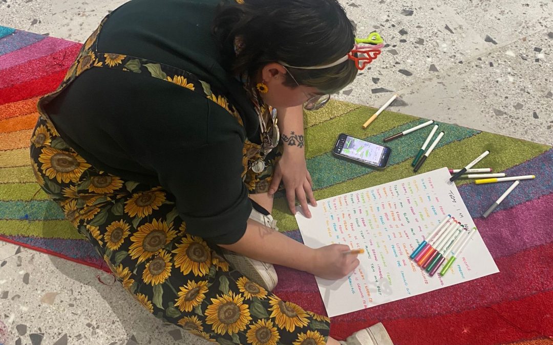 An image of Amy, in sunflower dungarees, writing a poem on a rainbow rug.