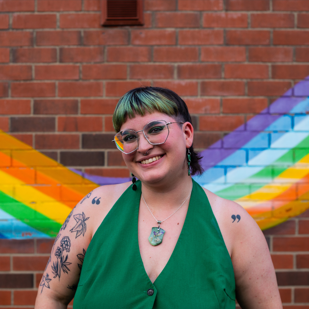 Amy, wearing a green suit, in front of a rainbow mural.